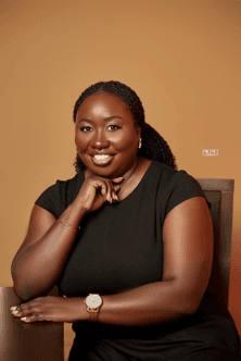 Studio portrait of Emma Wenani seated in a black dress, smiling with her chin on her hand